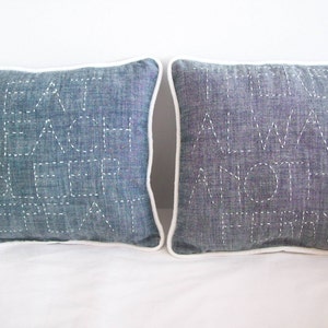 Op de afbeelding: Twee blauwe denim kussens met witte stiksels. Het kussen aan de linkerkant zegt "Eat Beach Sleep Repeat" en het kussen aan de rechterkant zegt "There's Always Another Ferry".