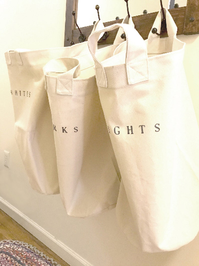 Pu&ograve; includere: Tre sacchi per la biancheria in tela bianco sporco appesi a una parete. Ogni sacco ha un manico ed &egrave; etichettato con una parola in lettere nere: "WHITES", "DARKS" e "LIGHTS".