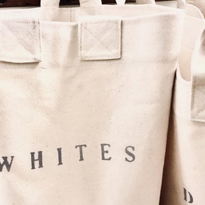 Pu&ograve; includere: Borsa per la biancheria in tela bianco sporco con la scritta "WHITES" stampata in grigio. La borsa ha una forma rettangolare con la parte superiore e le maniglie rinforzate. La borsa &egrave; appesa a un portabiancheria in legno.