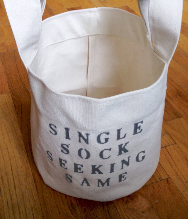 K&ouml;nnte beinhalten: Ein wei&szlig;er Canvas-W&auml;schekorb mit Griffen und dem aufgedruckten Text "SINGLE SOCK SEEKING SAME" in Grau.