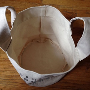 K&ouml;nnte beinhalten: Eine wei&szlig;e Canvas-Tragetasche mit zwei Henkeln. Die Tasche ist leer und das Innere ist sichtbar. Die Tasche hat schwarze Beschriftung auf dem Boden.