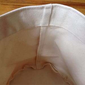 K&ouml;nnte beinhalten: Eine wei&szlig;e Canvas-Tragetasche mit einer Naht in der Mitte. Die Tasche ist leer und das Innere ist sichtbar.