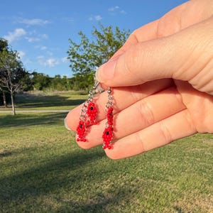 May include: A pair of handmade red beaded earrings with a fish design. The earrings are held up against a background of green grass and a blue sky. The earrings have a silver chain and hook.