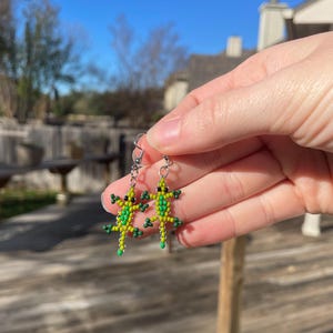 May include: A pair of beaded lizard earrings. The earrings feature a green and yellow color scheme with black accents. The lizards have small beaded legs and tails, and are suspended from silver-colored ear wires. The earrings are held up against a blurred outdoor background.