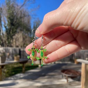 May include: A pair of beaded lizard earrings. Each earring features a green lizard with black eyes and small limbs. The lizards are made of small green beads and are suspended from silver-colored ear wires.