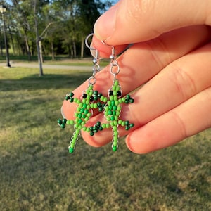 May include: A pair of beaded lizard earrings. The earrings are green with black eyes and small green beaded feet. The earrings are held up by silver-colored hooks. The background is a blurred view of a park with trees and grass.