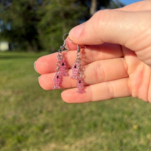 May include: Pink beaded fish earrings with black accents. The earrings are dangling from silver-colored hooks and chains. The fish design is made of small, translucent pink beads, creating a textured appearance. The earrings are held up by a hand.