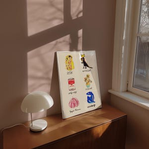 Puede incluir: Un cuadro enmarcado con seis ilustraciones coloridas, incluyendo un dinosaurio con corona, una lata de sopa y una calabaza rosa con lunares. La impresi&oacute;n est&aacute; sobre una superficie de madera junto a una l&aacute;mpara blanca con forma de hongo.