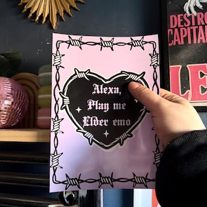 Può includere: Un adesivo rosa con un design a forma di cuore nero e un bordo di filo spinato. Il testo "Alexa, Play me Elder emo" è stampato sul cuore. L'adesivo è tenuto davanti a un muro blu scuro.