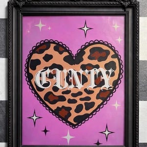 May include: Framed artwork with a pink backdrop, a leopard print heart, and the word "CUNTY" in white. The black frame has decorative elements, and the background includes star motifs. The artwork is framed in a black frame.