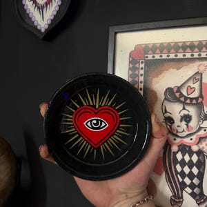 May include: A round black tray featuring a central graphic of a red heart with a white and black eye inside. The heart is surrounded by gold radiating lines. The tray has a smooth, dark finish and a circular shape with a raised rim.