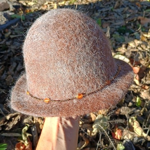 May include: A brown felted hat with a rounded crown and a short brim. The hat features a thin, dark beaded band encircling the base of the crown, accented with small, translucent orange beads spaced evenly along the wire.