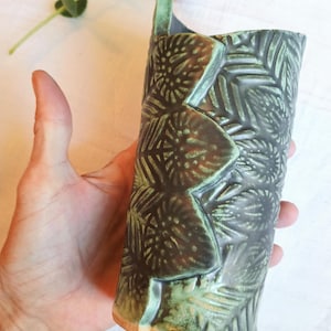 Può includere: Un vaso cilindrico in ceramica con una base grigio antracite scuro e un motivo a foglie in rilievo evidenziato da uno smalto verde menta chiaro. Il contenitore presenta un bordo irregolare e una base in argilla color terracotta visibile.
