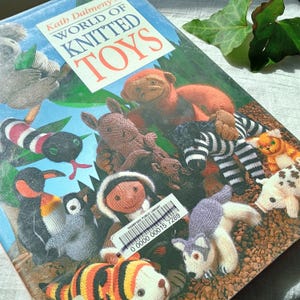 May include: A book cover titled "World of Knitted Toys" by Kath Dalmeny, featuring various knitted animal toys. The toys include a monkey, zebra, penguin, and a husky. The cover is blue with green and brown accents.
