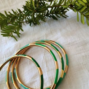 May include: A collection of gold bangles featuring green enamel detailing. The bangles display a straightforward design with alternating gold and green segments. A sprig of green foliage is visible in the background.