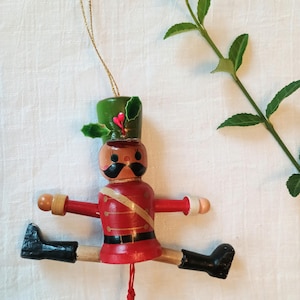 May include: A wooden nutcracker ornament featuring a red jacket with gold trim, a green hat with holly leaves, and black boots. The figure has a black mustache and is suspended by a gold string, with a red pull string attached to the bottom.