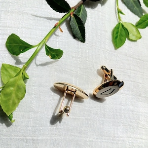 May include: A pair of gold-toned cufflinks with an oval design. One cufflink features a black and white arrow design. The cufflinks are set against a white background with green leaves.