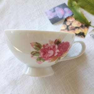 May include: A white teacup with a floral rose design in shades of pink and green. The cup has a handle and sits on a small pedestal base. The teacup is on a white surface.