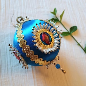 May include: A spherical blue satin ornament decorated with gold metallic lace trim, a central red diamond-shaped gem, and a radiating border of small white beads. The ornament features a silver hanging loop at the top and a small dangling blue bead.