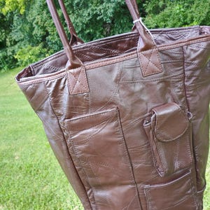 Puede incluir: Bolso tote de cuero marrón con diseño de patchwork. El bolso tiene dos asas largas y varios bolsillos exteriores. El bolso está hecho de cuero marrón y tiene forma rectangular. El bolso se muestra sobre un fondo verde.