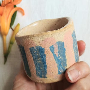 May include: A small, cylindrical ceramic cup with a speckled beige base featuring vertical, hand-painted brushstrokes in alternating shades of soft pink and vibrant blue. The cup has a textured, matte finish and a slightly uneven, organic rim.