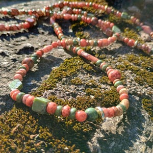 May include: A handmade necklace featuring a mix of red, green, and brown beads. The necklace includes various shapes and sizes of beads, including round, square, and cylindrical. The beads are strung together with a silver-colored metal clasp.