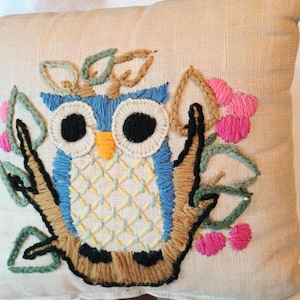 May include: A square beige throw pillow featuring an embroidered blue owl with large black eyes and a yellow beak. The owl is perched on a brown branch surrounded by green leaves and small pink flowers, all rendered in textured embroidery thread.