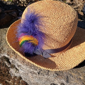 May include: A woven straw hat with a dark ribbon and a cluster of purple and rainbow-coloured feathers. The hat is resting on a stone surface, likely handmade.