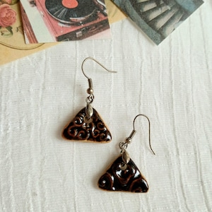 May include: A pair of dangle earrings featuring triangular ceramic charms with a glossy dark brown glaze and embossed swirling patterns. The charms are suspended from silver-toned metal fishhook wires with small decorative metal loops at the top.