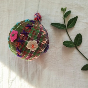 May include: A spherical fabric ornament featuring a green plaid pattern with decorative hand-stitched embroidery in purple, light blue, and pink. The ornament includes small felt flower appliqués and a thin silver wire loop for hanging purposes.