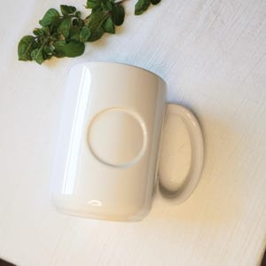 May include: A white ceramic mug with a large, rounded handle and a circular indentation on the side. The mug is set against a white background with a sprig of green leaves.