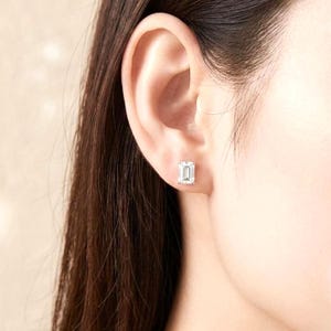 May include: A close-up of an ear with a square-cut, clear gemstone stud earring. The earring is set in a silver or white metal. The person's skin is fair, and their hair is dark brown.