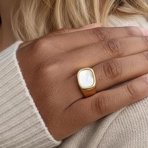 May include: A gold-colored ring with a square, white mother-of-pearl inlay. The ring is worn on a finger, and the design is simple and elegant. The ring is a piece of jewelry.