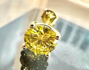 May include: Close-up of a yellow gemstone stud earring with a gold setting. The round-cut gemstone is a vibrant yellow color and reflects light. The earring is set on a reflective surface.