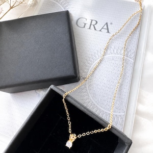 May include: A gold-toned necklace featuring a single round clear gemstone pendant in a four-prong setting. The delicate chain is displayed partially inside a black jewelry box next to a GRA certificate document with a circular sunburst graphic.