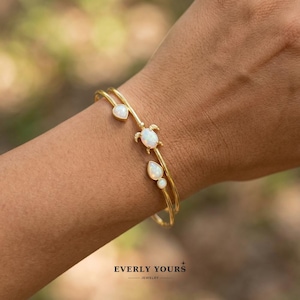 Puede incluir: Pulsera dorada con tres detalles de ópalo y un pequeño dije de tortuga. La pulsera presenta un ópalo en forma de corazón, un ópalo en forma de lágrima y un ópalo en forma de tortuga. La pulsera se lleva en el brazo de una persona.