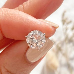 May include: A round, brilliant-cut clear gemstone set in a silver-toned metal stud earring. The stone features multiple facets that reflect light, creating a sparkling appearance. The setting includes small prongs securing the stone in place.