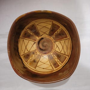 May include: A handmade ceramic bowl with a square shape and a circular design in the center. The bowl features a brown exterior and a tan interior with geometric patterns in brown and tan. The center has a spiral design.