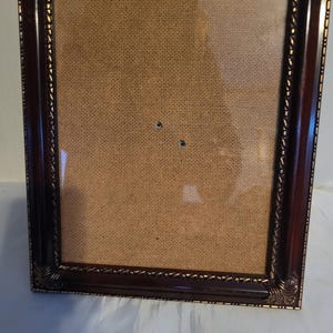 May include: A rectangular picture frame with a dark brown finish and ornate gold-colored detailing. The frame has a glass front and a tan backing. The corners of the frame have decorative scrollwork.