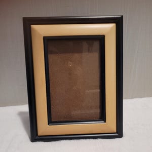 May include: A rectangular picture frame with a black outer frame and a light tan inner frame. The frame has a brown backing and a glass or clear plastic cover. The frame is designed to hold a photograph or artwork.