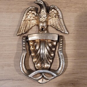 May include: A vintage door knocker featuring a detailed eagle with outstretched wings above a decorative handle. The eagle and handle have a gold and silver finish, mounted on a wooden surface. The door knocker is a decorative hardware piece.