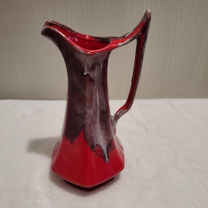 May include: A red and brown ceramic pitcher with a handle. The pitcher has a unique shape with a wide base and a narrow neck. The interior is red, and the exterior has a glossy finish with a blend of red, brown, and white hues.