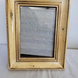 May include: A rectangular picture frame with a gold-colored trim and a cream-colored border. The frame has a distressed finish, with visible imperfections. The frame is empty, with a gray interior.