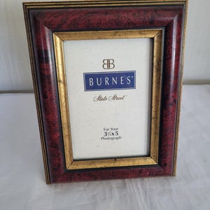 May include: A rectangular photo frame with a burgundy and gold finish. The frame has a gold inner border and a burgundy outer border with a wood grain pattern. The frame is designed for a 3.5 x 5 inch photograph. The brand name "BURNES" is visible.