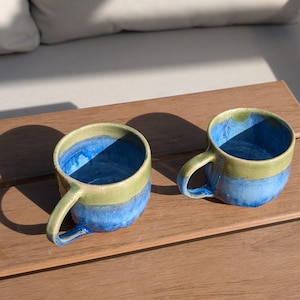 Puede incluir: Dos tazas de cerámica hechas a mano. Cada taza tiene un interior azul, una banda verde claro alrededor del borde y una base azul. Las tazas están sobre una superficie de madera, con sombras.