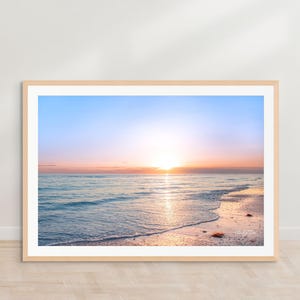 May include: A framed photograph of a sunset over the ocean. The sky is a gradient of blue, pink, and orange, and the sun is setting in the distance. The waves are crashing on the shore, and the sand is white.