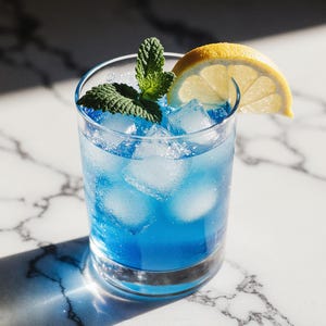 Può includere: Un cocktail blu rinfrescante in un bicchiere trasparente, pieno di cubetti di ghiaccio, guarnito con un rametto di menta e una fetta di limone. La bevanda è su una superficie di marmo bianco.