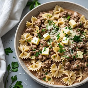 Puede incluir: Un bol blanco lleno de pasta de lazo, carne molida y mantequilla derretida. La pasta está cubierta con perejil fresco y queso rallado.