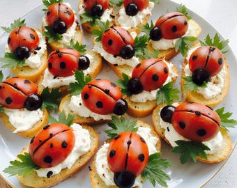 May include: A plate of appetizers featuring ladybug-shaped food. Each ladybug is made from a halved cherry tomato with black olive spots and a black olive head, atop a bed of white cream cheese and parsley on toasted bread slices.