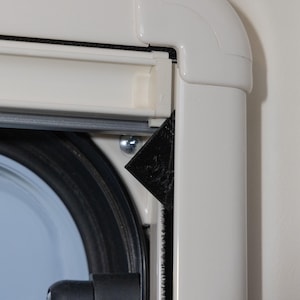 Puede incluir: Primer plano de un marco de ventana blanco con una ventana redondeada. El marco tiene un sello de goma negro y una pieza de esquina negra. Se ve un tornillo plateado. El fondo es una pared clara.